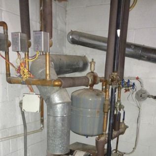 Complex industrial pipes, valves, and equipment, including a gray pressure tank, against a white wall.