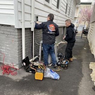 Two people working on a unit next to a house. Tools, electrical cords on ground.
