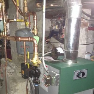 Green boiler with copper pipes and a silver chimney in a basement.