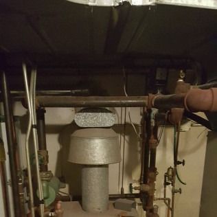 Overhead view of pipes and equipment in a basement. A metal vent has a stone on top of it.