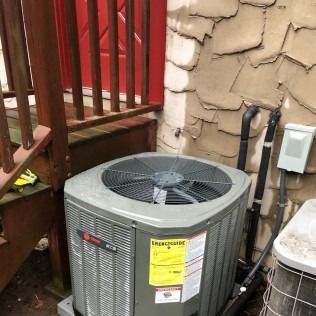 Gray air conditioner unit next to a brown deck and tan textured wall.