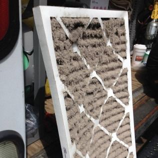 A heavily soiled air filter, showing thick dust accumulation and some damage, held up outdoors.