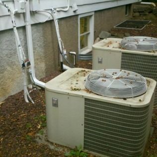 Two outdoor air conditioning units near a house. They are tan, with protective fan covers, and covered with debris.