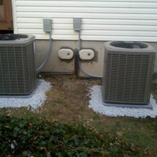 Two air conditioning units side-by-side against a house, on gravel base. Electrical boxes above.