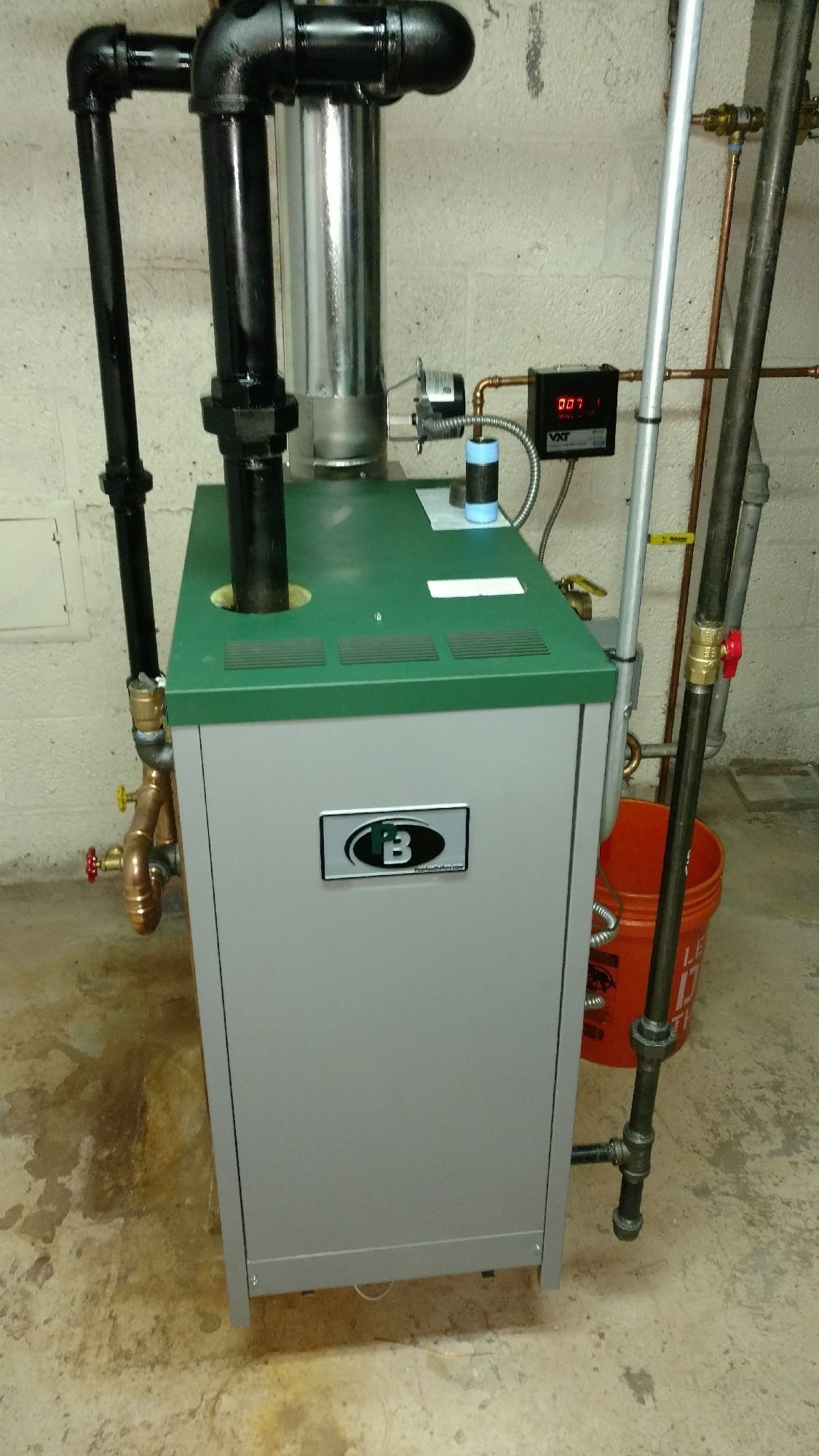 Gray and green furnace in a basement setting with black pipes.