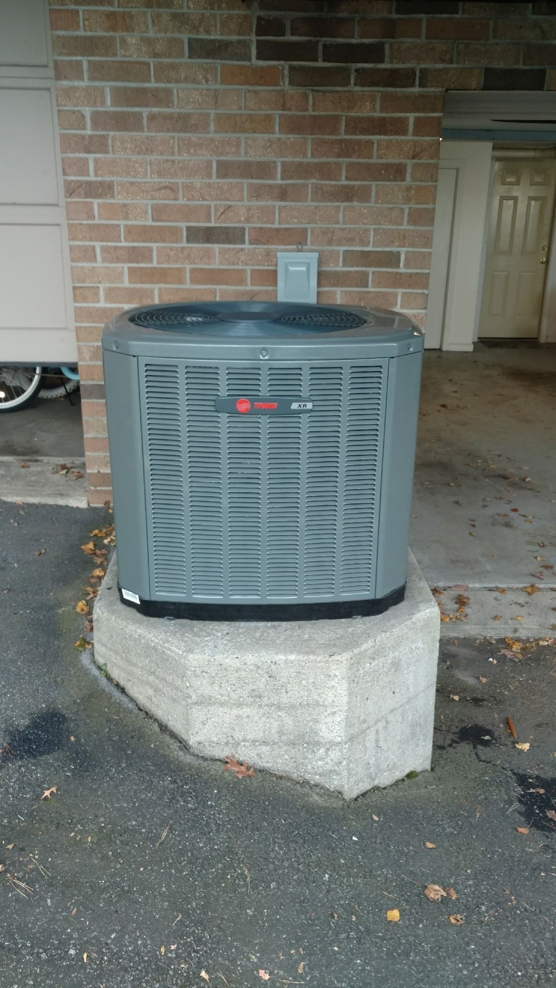 Gray air conditioning unit on a concrete pedestal against a brick wall.