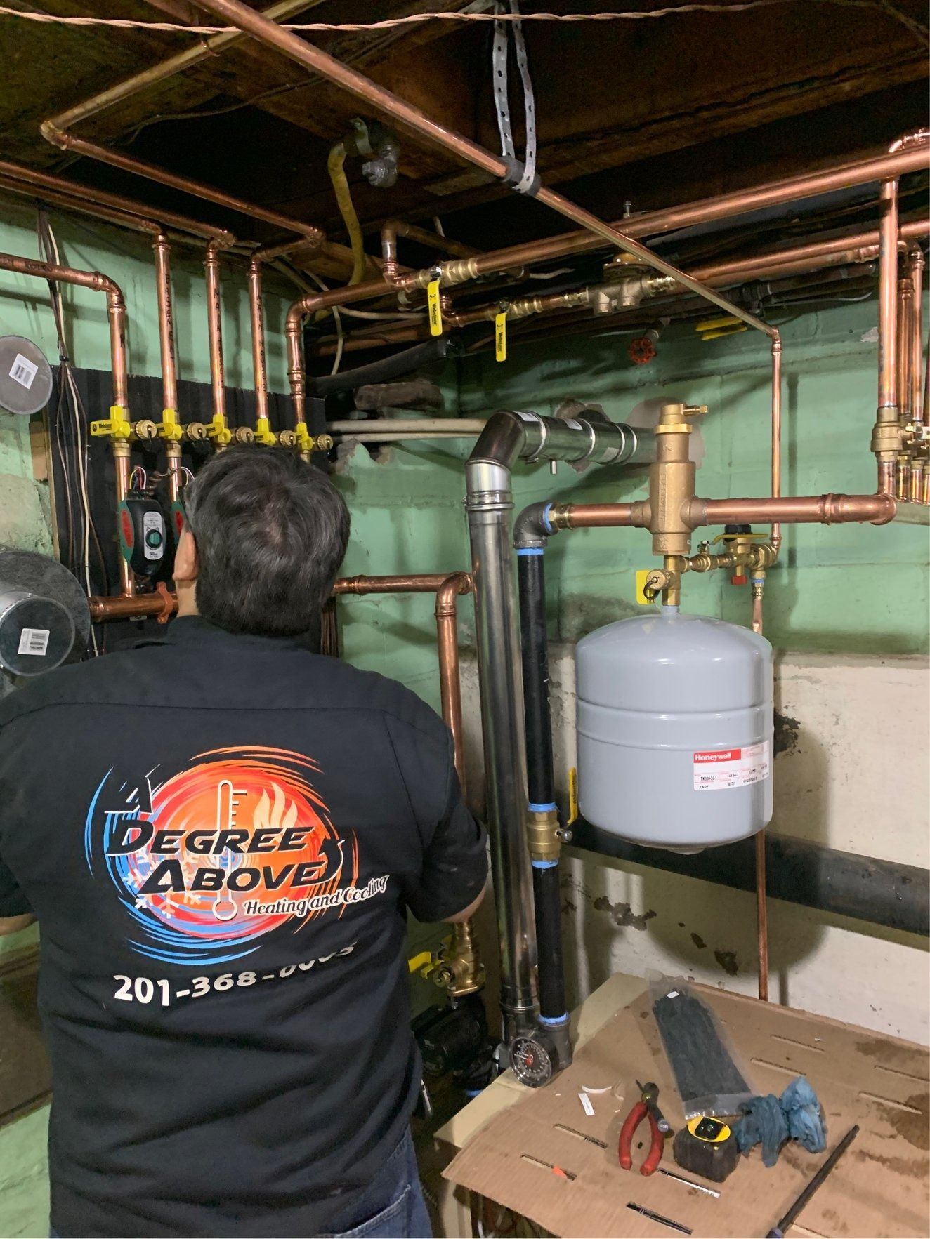 Plumber working on copper pipes in a basement.  He wears a black shirt with the company logo 