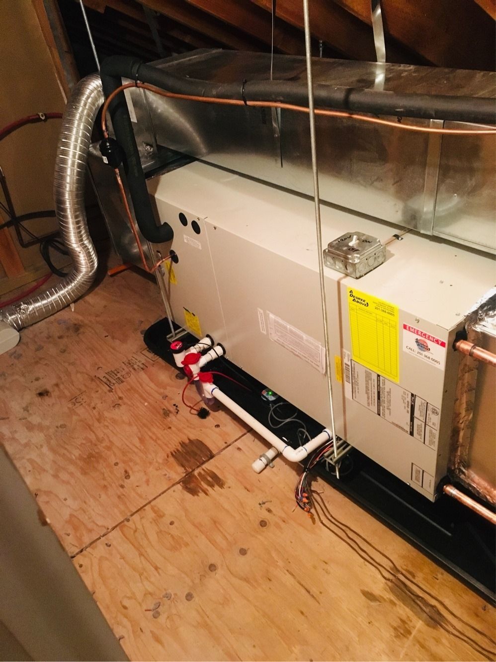 HVAC unit in an attic. Ductwork, piping, and electrical wires are visible. Beige unit on wooden floor.