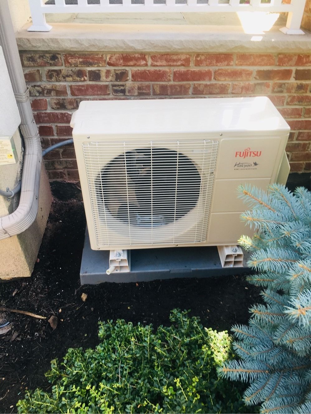 Fujitsu air conditioning unit on a concrete pad near a brick wall and bushes.