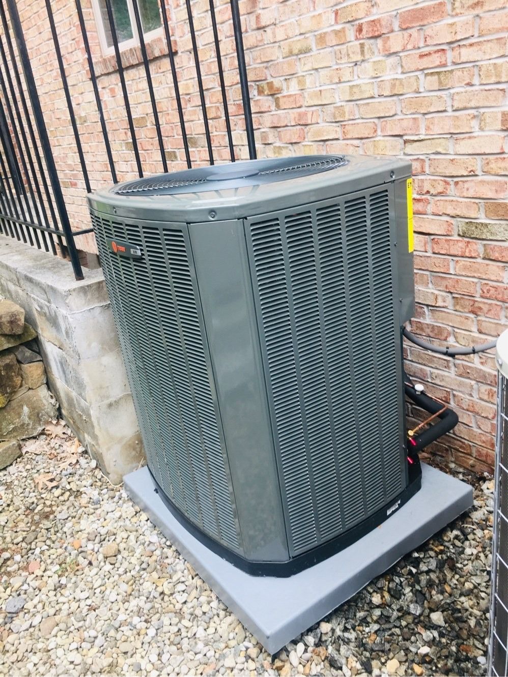 Gray Trane air conditioner unit on a concrete pad near a brick wall and black metal fence.
