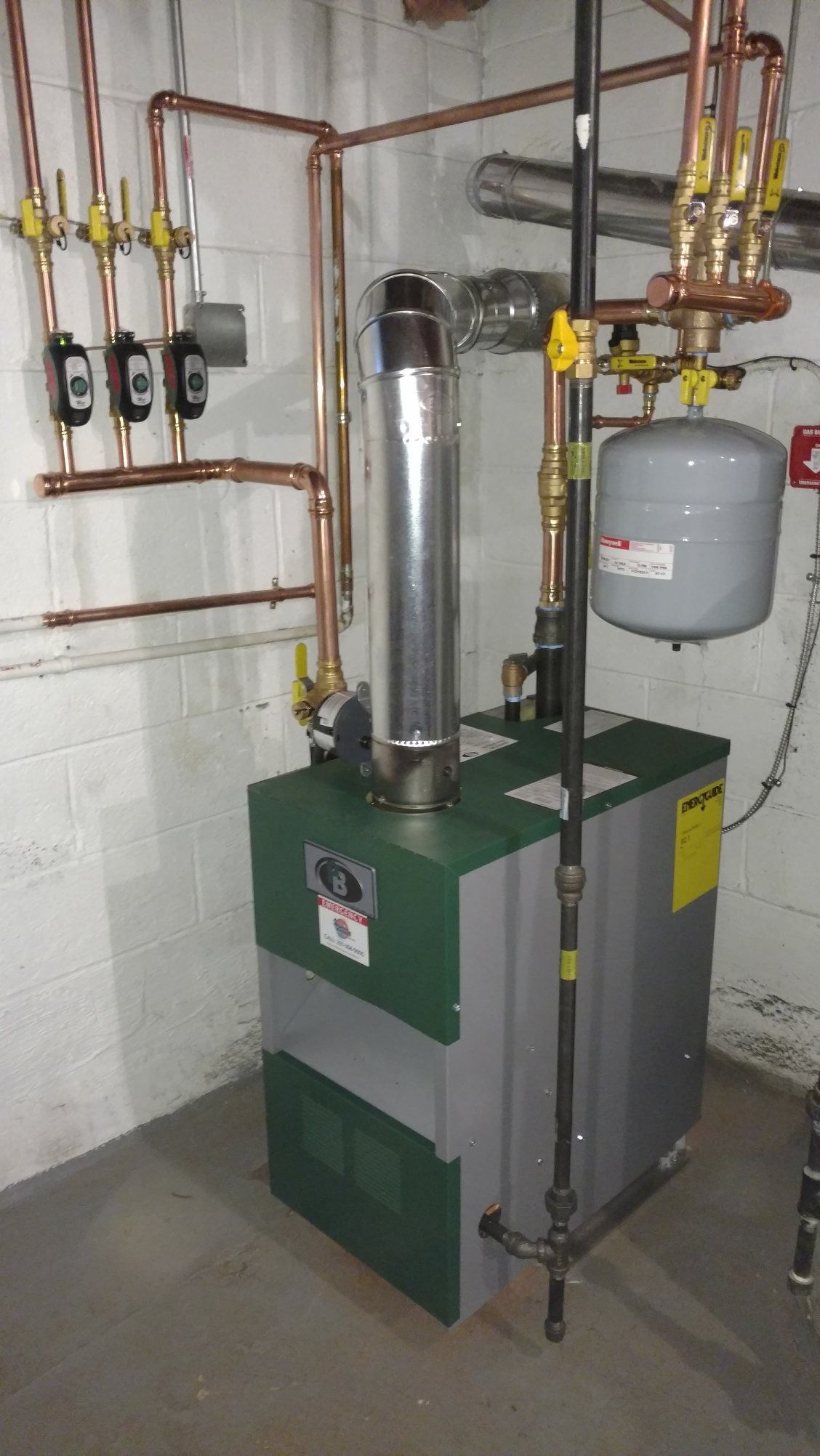 Green and gray furnace with copper pipes and chimney in a basement setting.