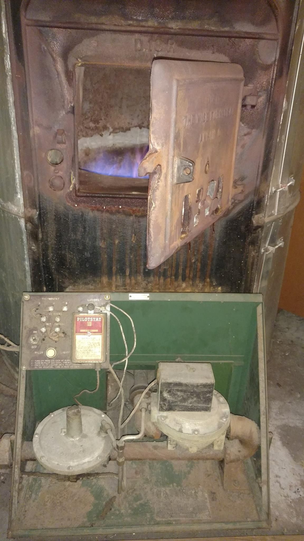 Old, rusty oil furnace with a blue flame visible. Green control panel below.