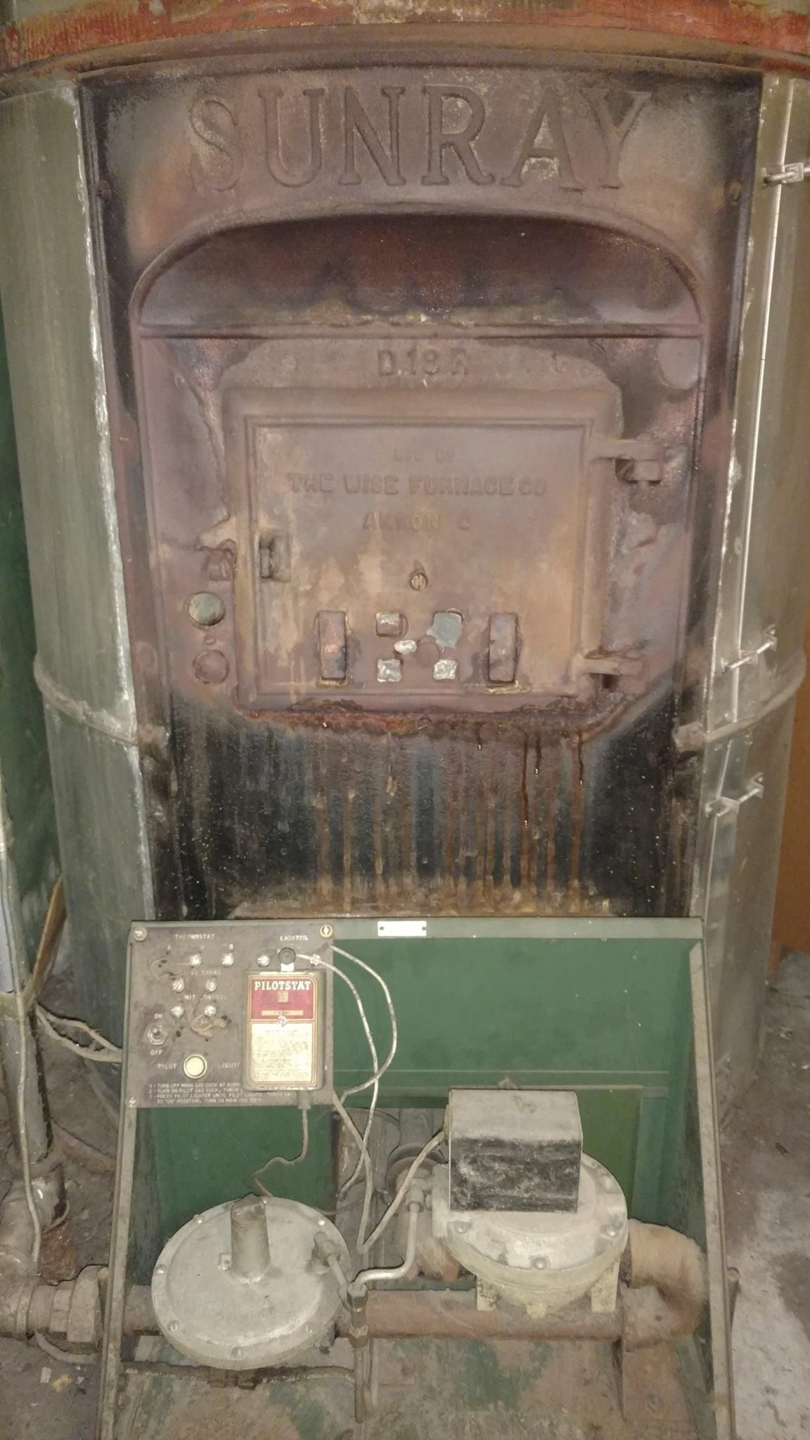 Old Sunray furnace with control panel, pipes, and fan. Metal and green components, possibly abandoned.