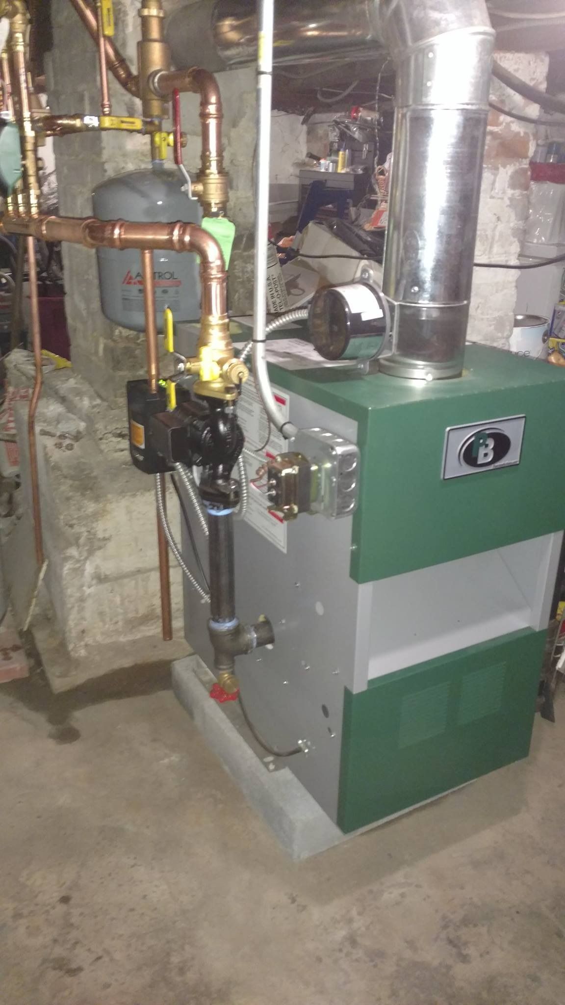 Green and gray boiler with copper pipes and a silver chimney in a basement.