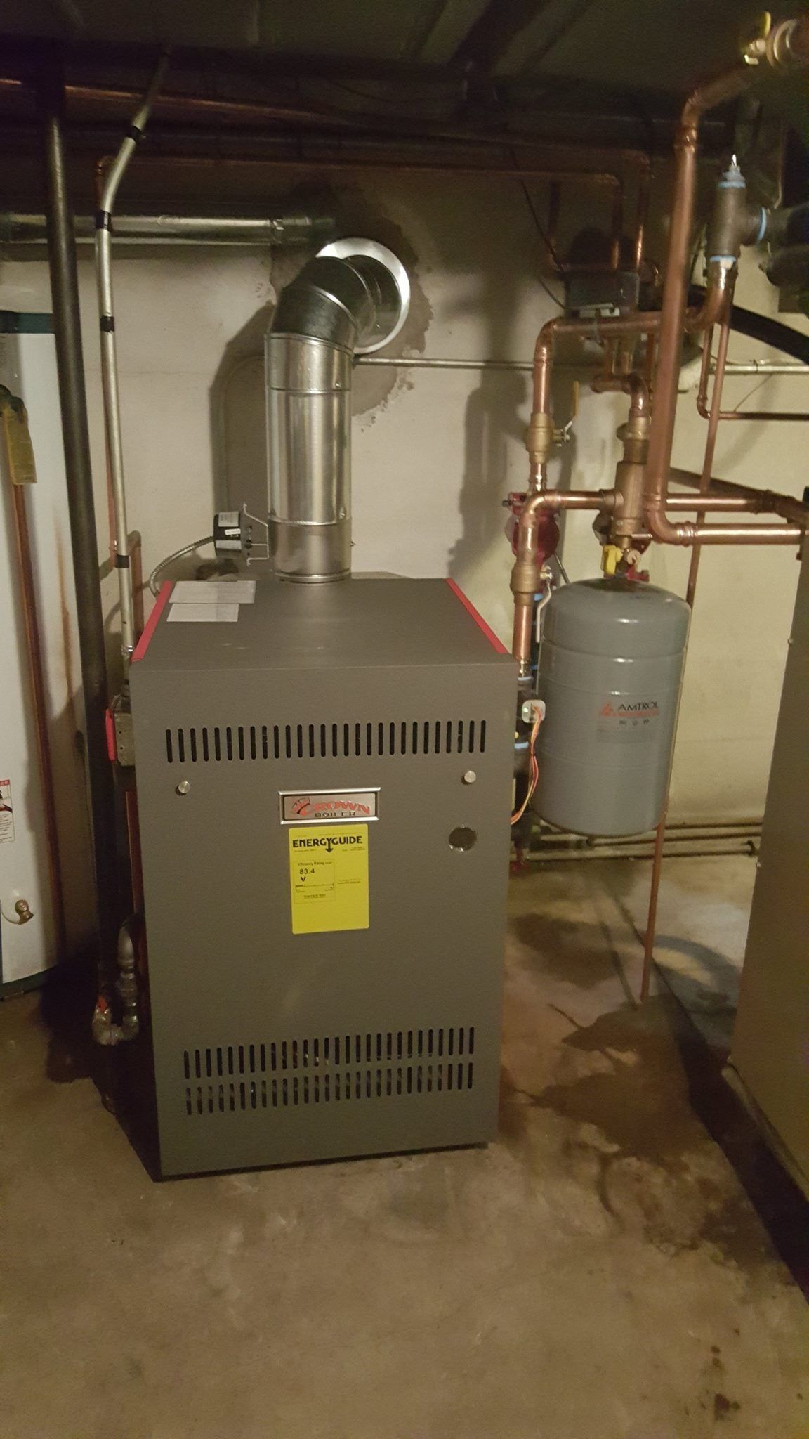 A gray boiler with a silver flue pipe and copper plumbing in a basement.