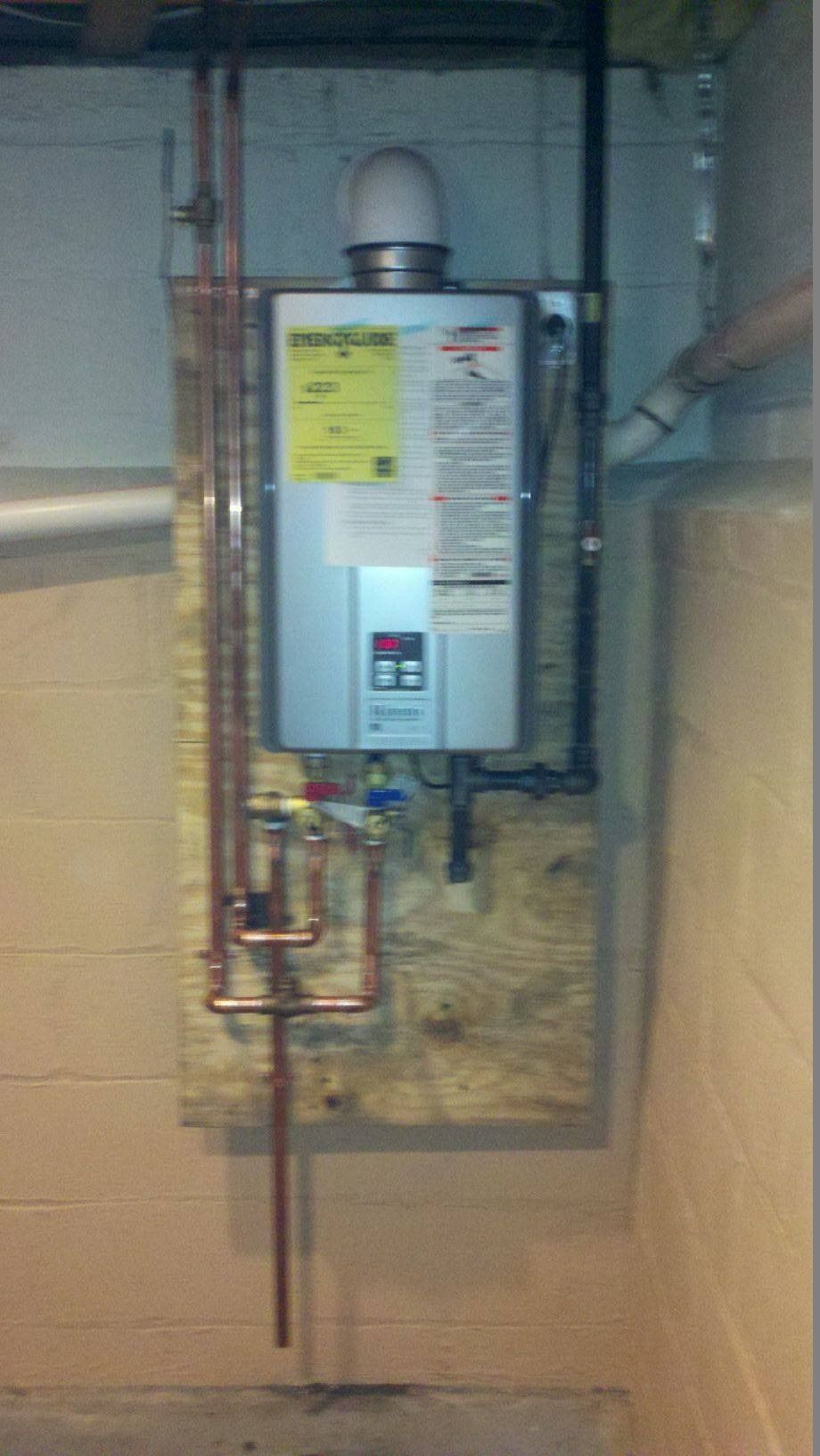 Tankless water heater installed on a plywood board in a basement. Copper pipes and black exhaust pipe are visible.