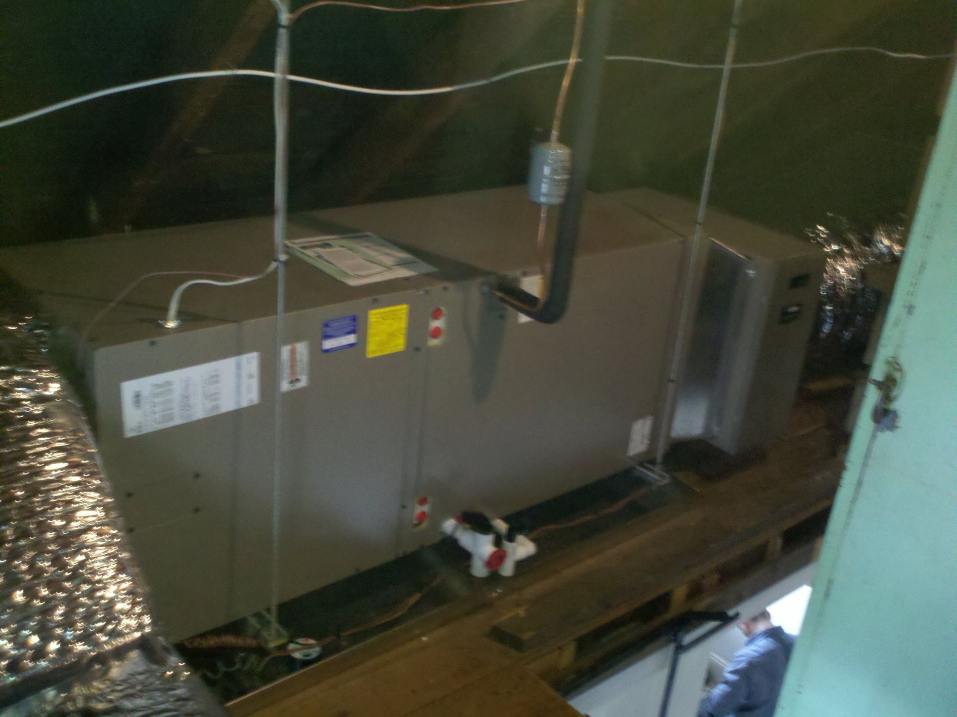 HVAC unit in attic, gray metal, with ductwork, electrical wiring. A person in blue is visible.