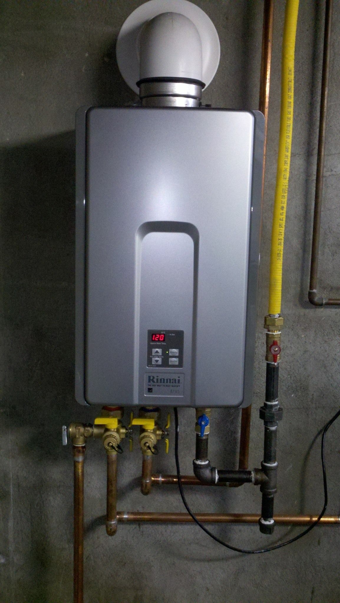Silver tankless water heater with vent pipe, installed on a concrete wall with gas and water lines.