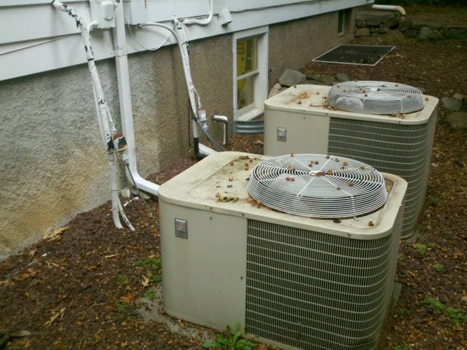 Two weathered air conditioning units outside a building with a window. Brown mulch and debris visible.