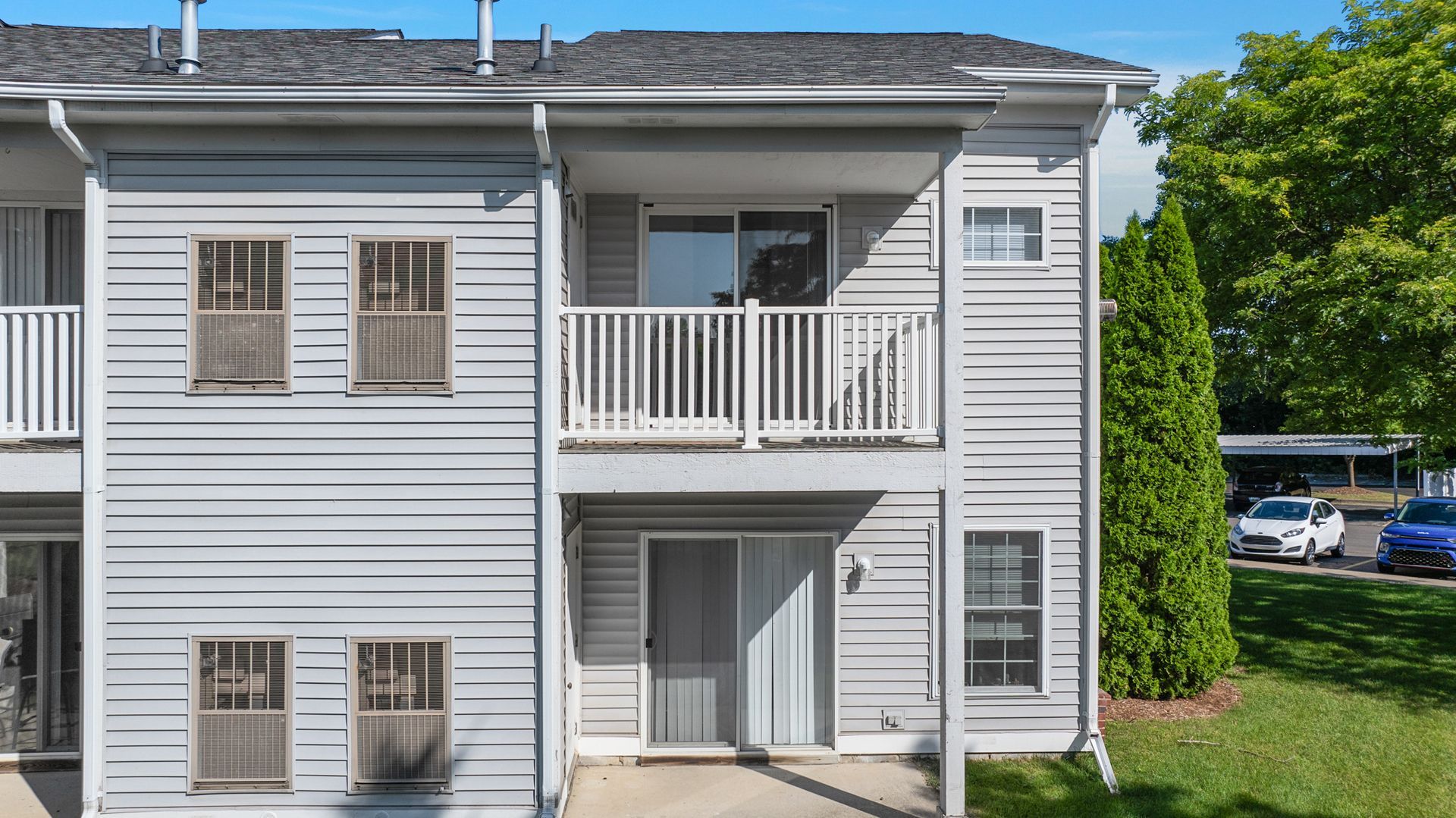 A white apartment building with a balcony and a parking lot in front of it.