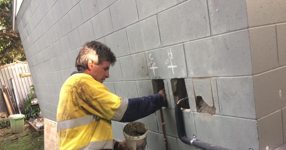 Man Working On Gray Cinder Block Wall Applying Cement To Pipe — David Talbot Plumbing in Cranbrook, QLD
