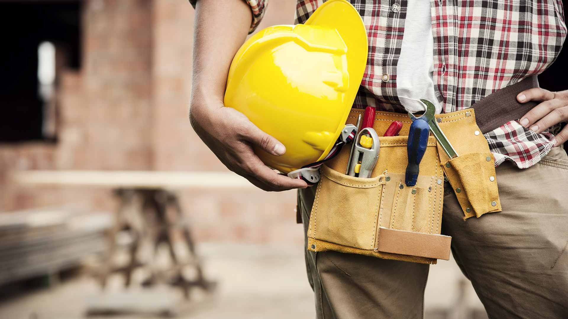 Construction Worker Holding Yellow Hard Hat With Tool Belt In Front Of Building — David Talbot Plumbing in Cranbrook, QLD