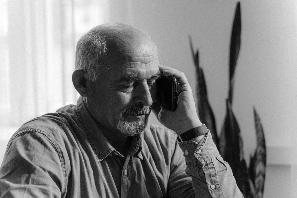 Man holding a phone to his ear, looking down thoughtfully. Indoors, near a plant.