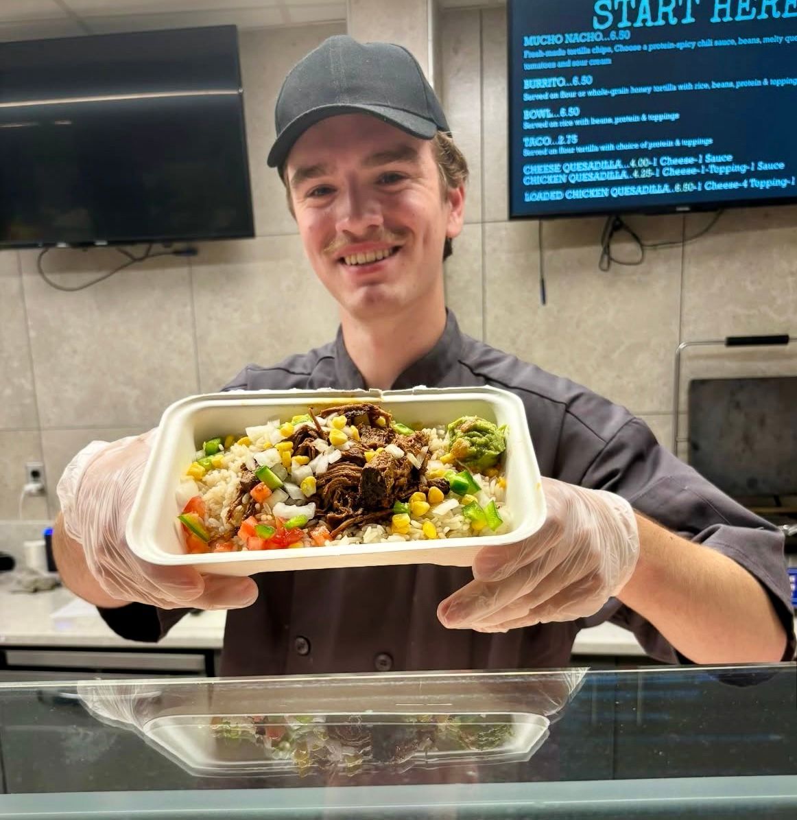 Man in a chef's uniform holding a food bowl in a restaurant. He is smiling and wearing gloves and a cap.