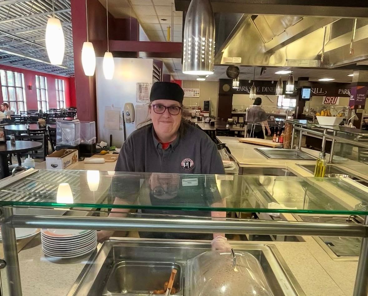 Person at a cafeteria counter, smiling. Commercial kitchen setting, food display case, overhead lighting.