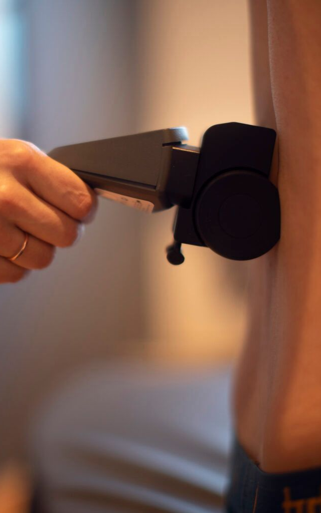 Mann gir ryggmassasje til en person som sitter i en massasjestol. Omgivelser: et terapirom.