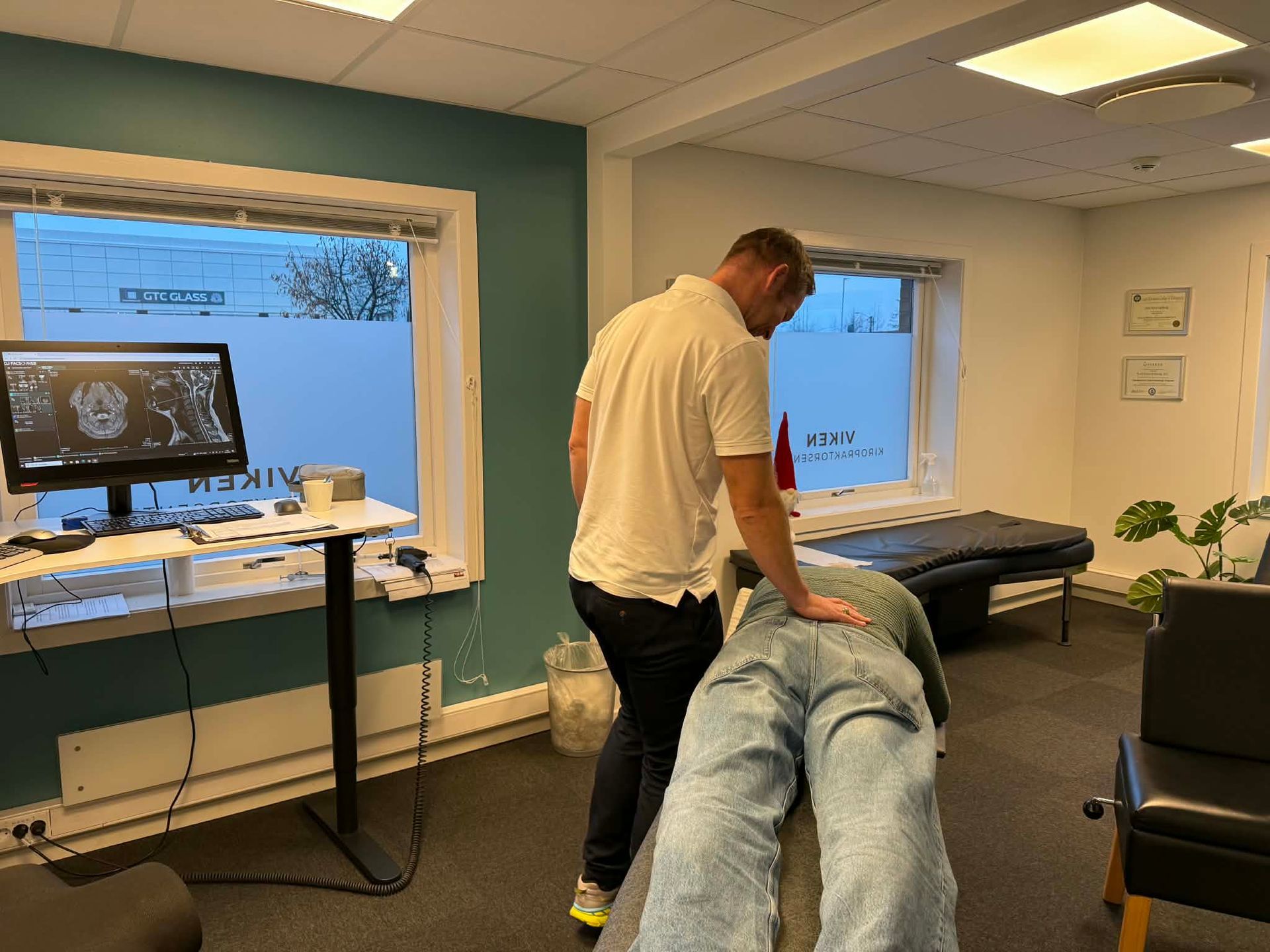 En helsearbeider undersøker en pasient som ligger med ansiktet ned på et bord; et innvendig rom med et vindu, en datamaskin og en stol.