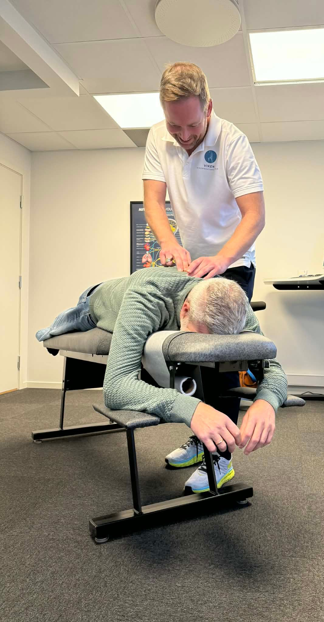 Mann gir ryggmassasje til en person som sitter i en massasjestol. Omgivelser: et terapirom.