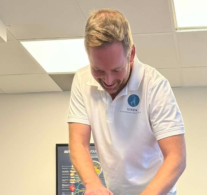 Mann i hvit poloskjorte, smilende, lent over et bord. Interiør, muligens en klinikk.