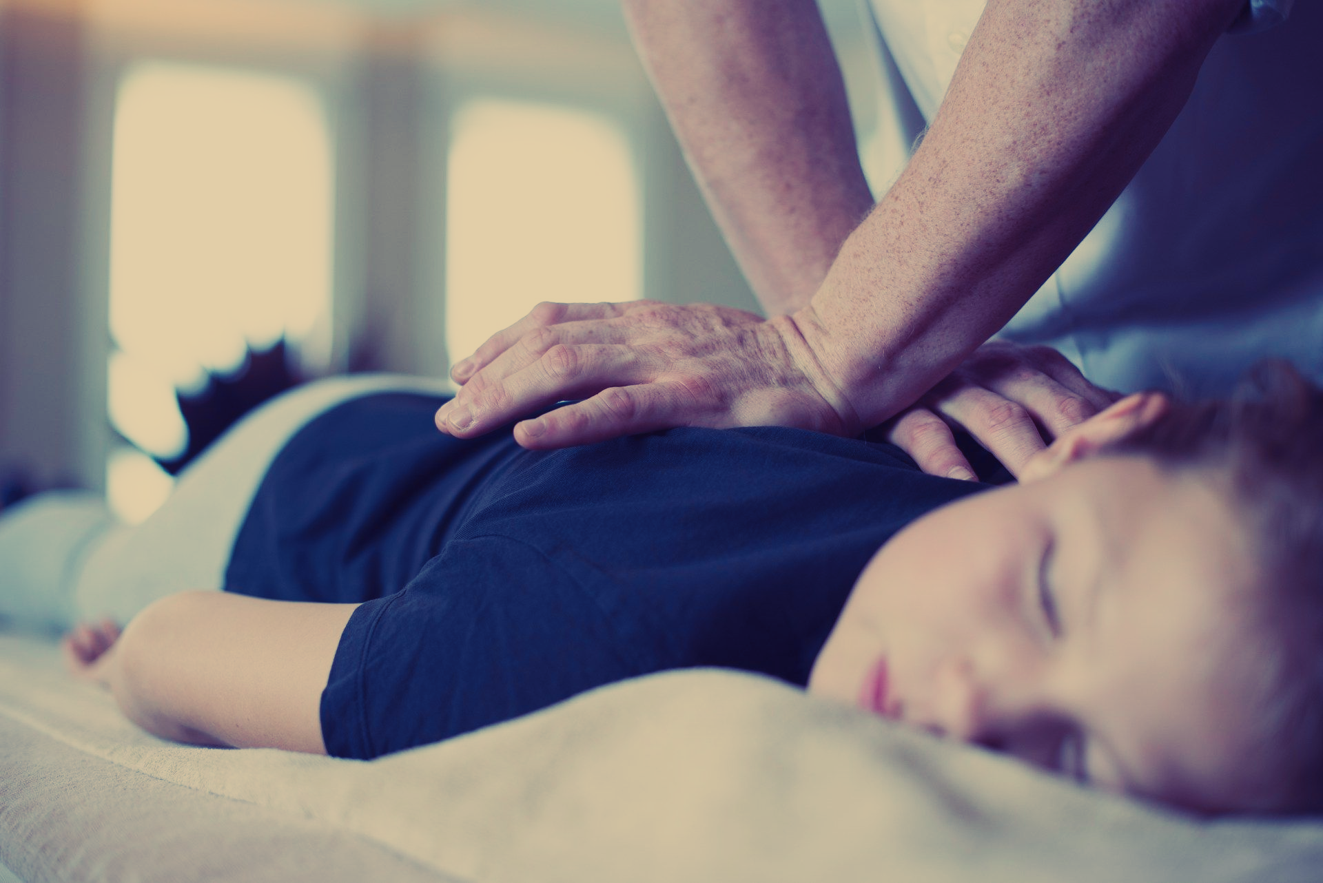En persons hender på et barns rygg, muligens under en massasje. Barnet ligger nede med lukkede øyne.
