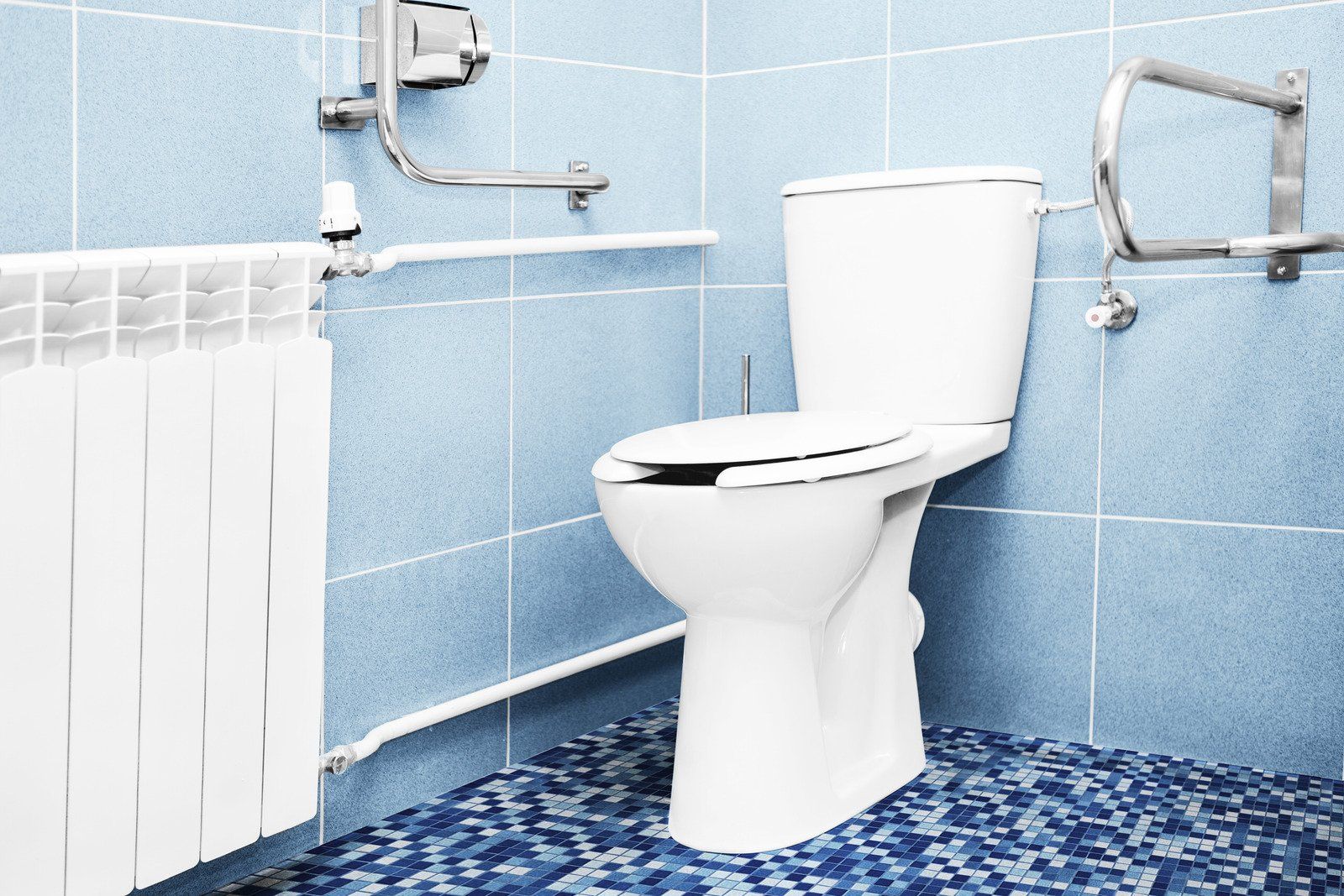 White toilet in a blue-tiled bathroom with grab bars and a radiator.