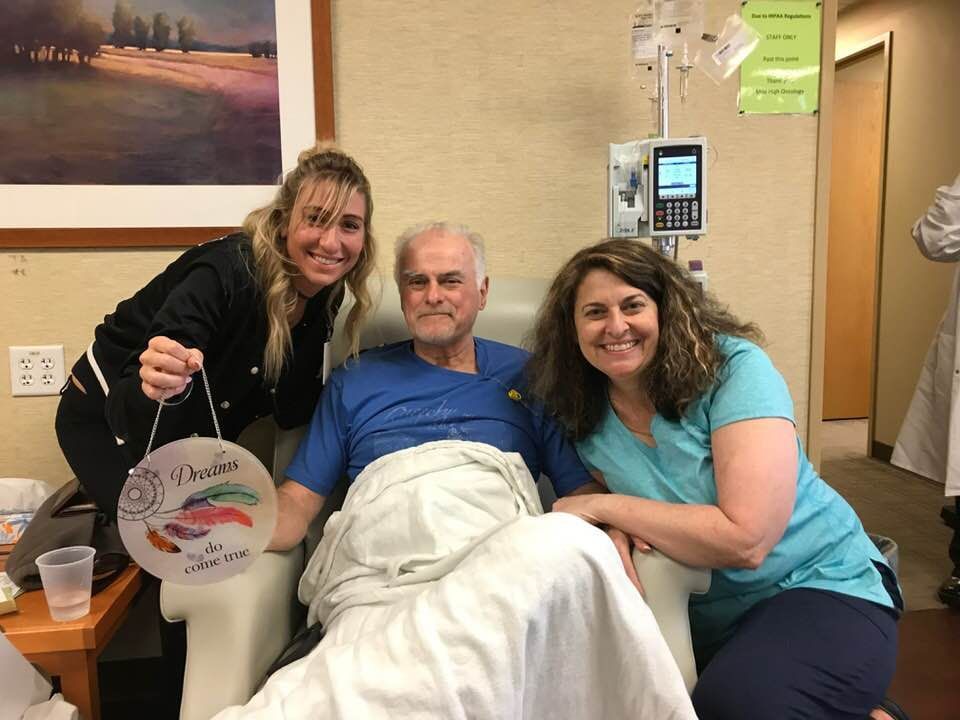 Man in chair with IV, smiling between two women in room. One woman holds a sign.