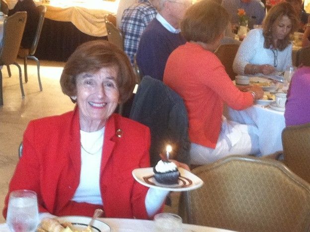 Elderly woman in red suit smiles, holding cupcake with lit candle at a restaurant table.