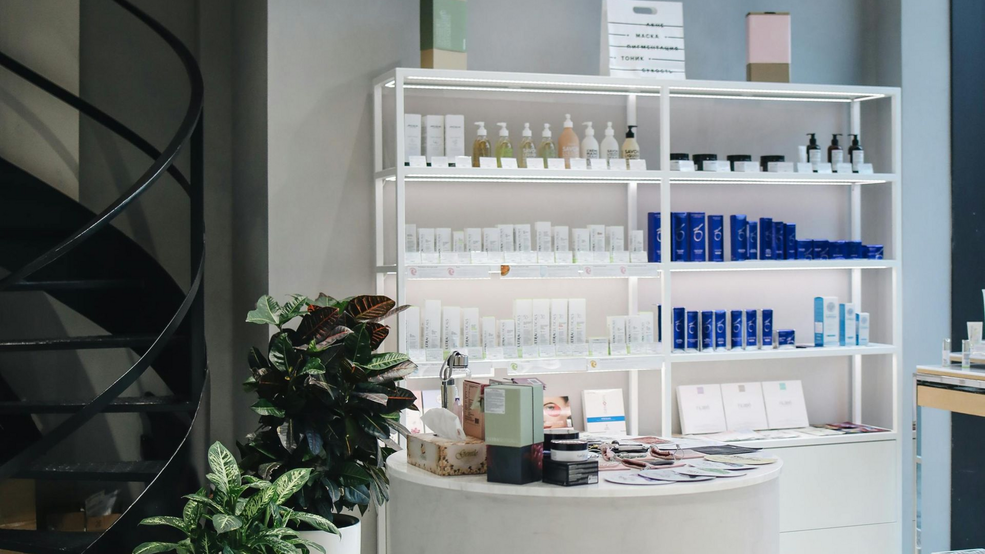Skincare products displayed on white shelves in a shop, with a spiral staircase and a round table with brochures.