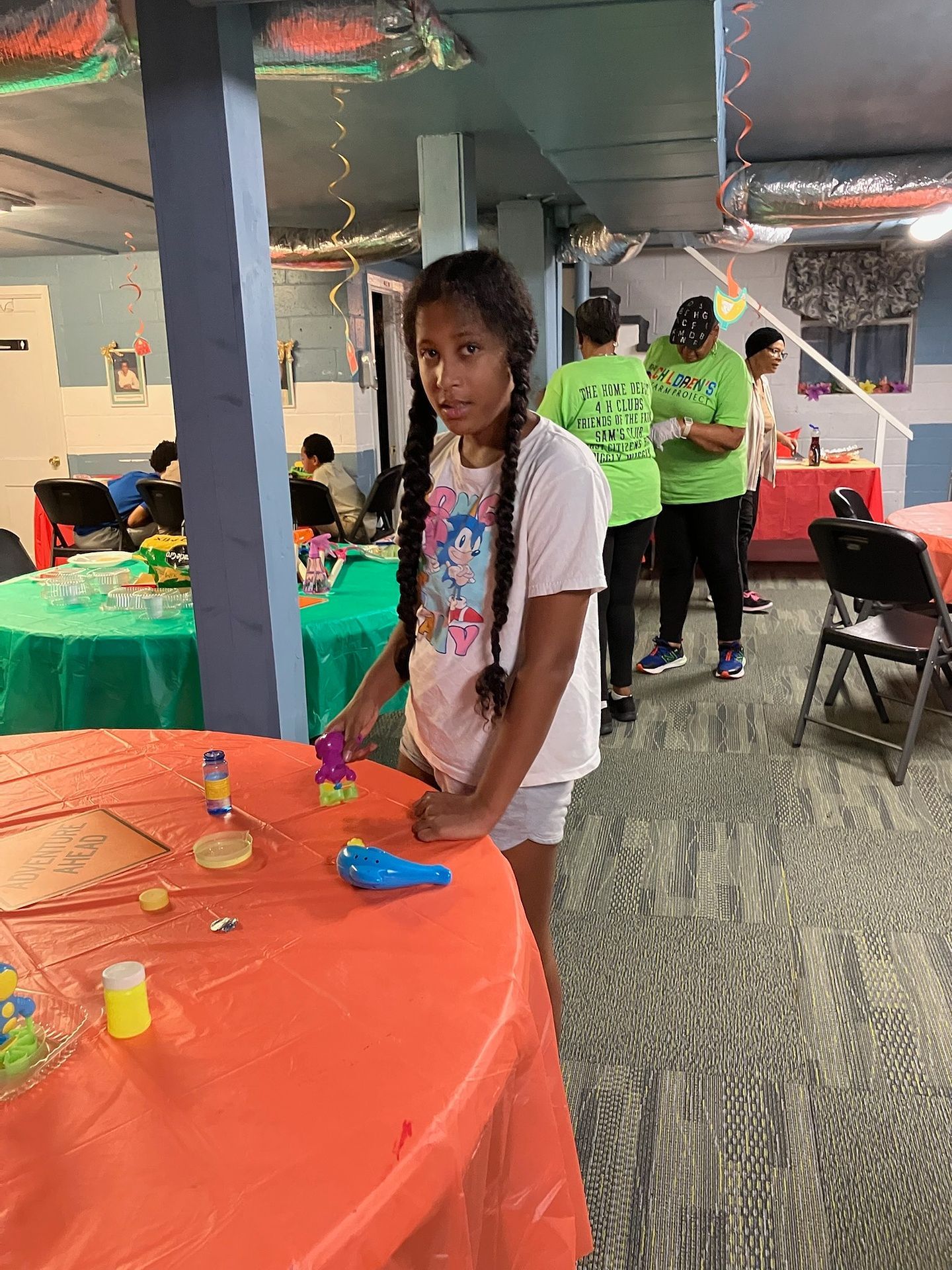 A person in a white T-shirt with braids stands by an orange table in a room with other people, tables, and decorations.