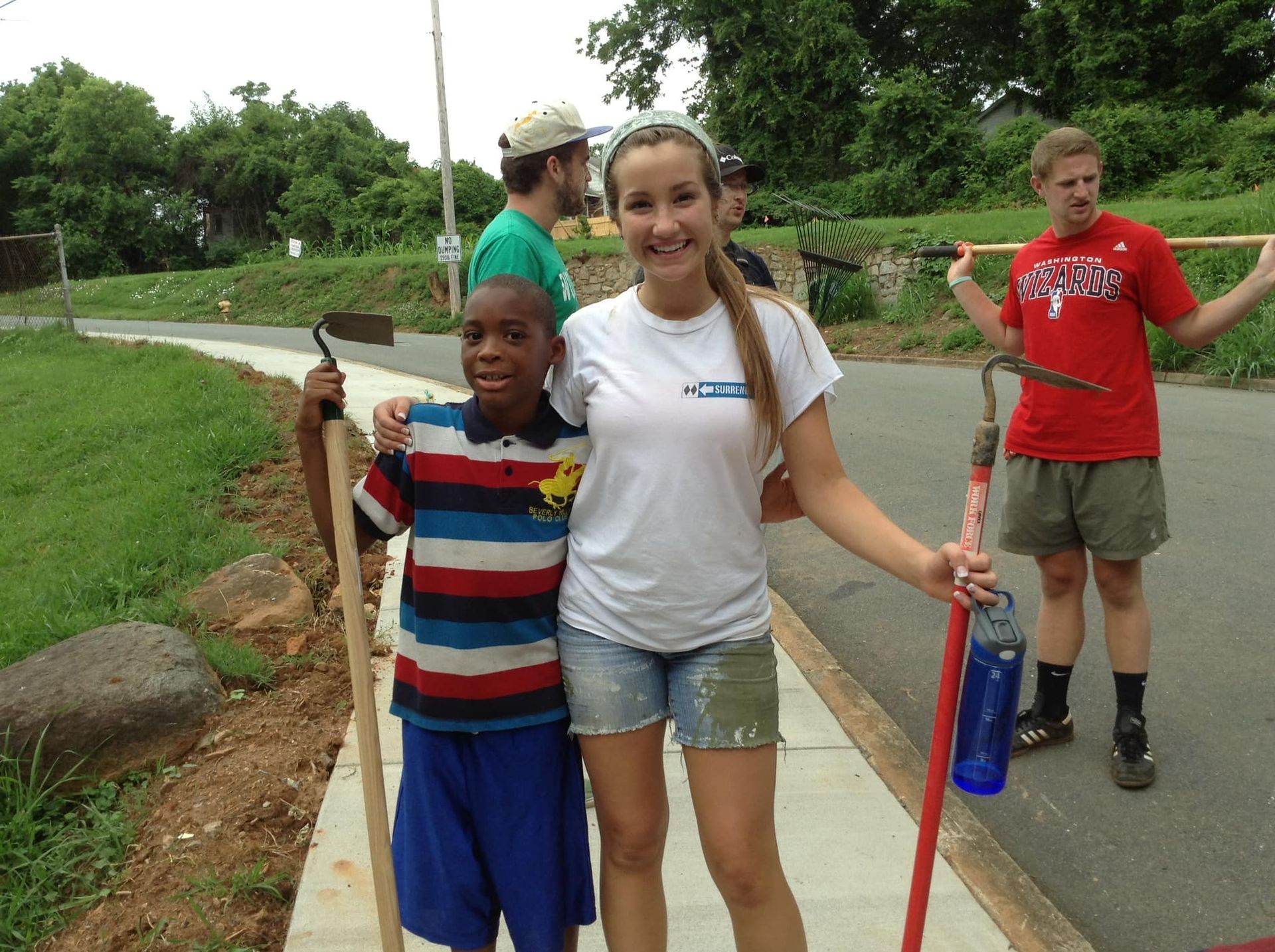 A group of volunteers posing outdoors on a sidewalk, some holding gardening tools, during a community service project.