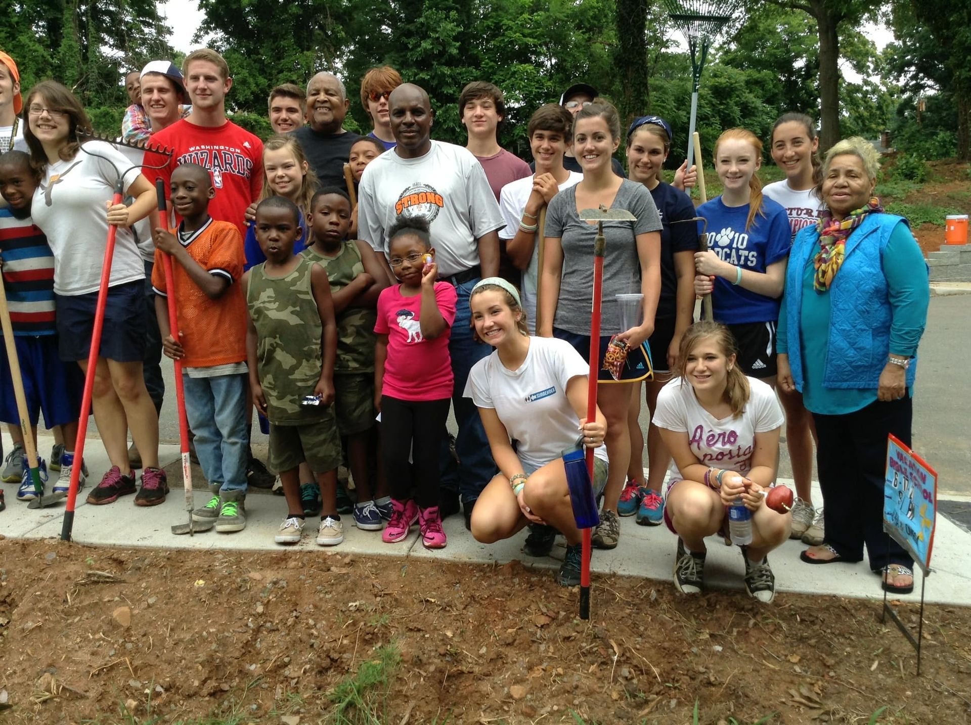 A diverse group of people posing outdoors with gardening tools, gathered in front of a freshly prepared soil bed.