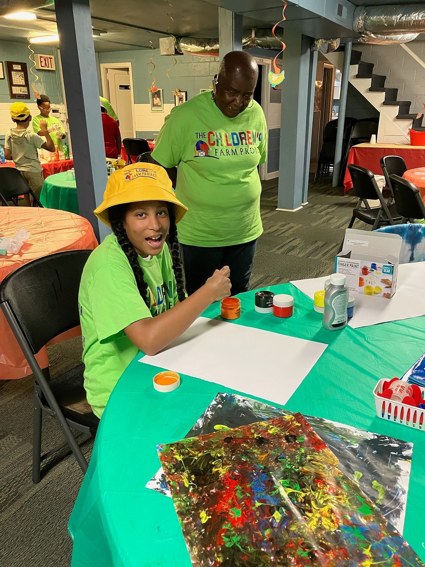 A child in a yellow bucket hat and green shirt painting on a large sheet of paper with an adult watching at a table.