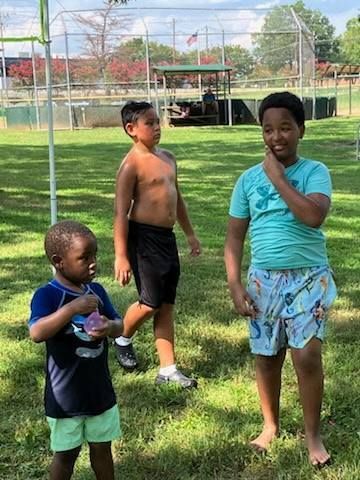 Three children in swimwear stand on a grassy field near a fenced baseball backstop on a sunny day.