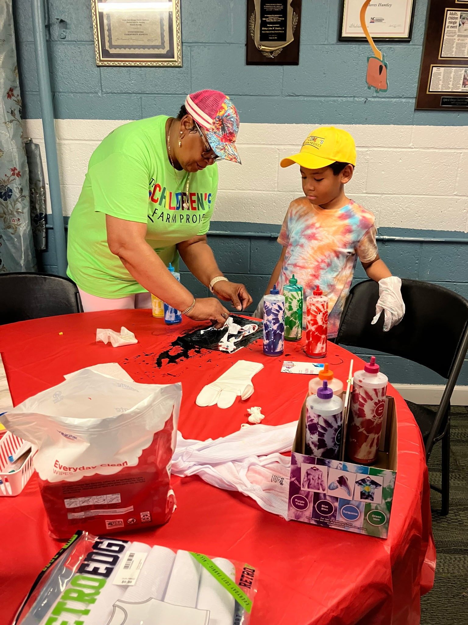 An adult and a child apply colorful dye from bottles onto a project on a red-covered table at an indoor event.