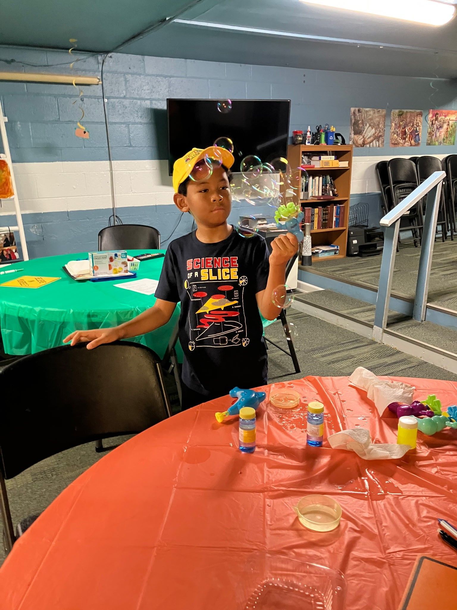 A person in a yellow cap and graphic t-shirt holds a bubble wand in an indoor activity room with colorful tables.