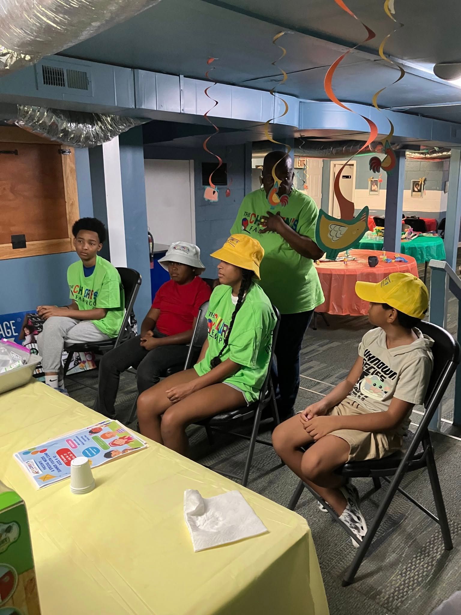 A group in neon green shirts and hats listens to an instructor in a basement space with tables and festive decorations.