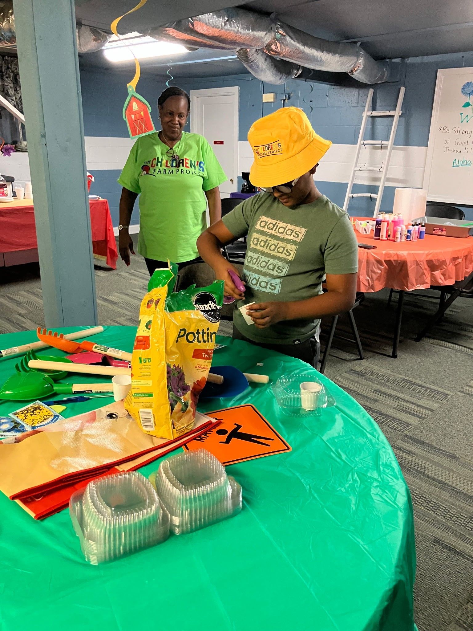 A person in a yellow hat works on a craft at a green-covered table while another person stands and watches nearby.
