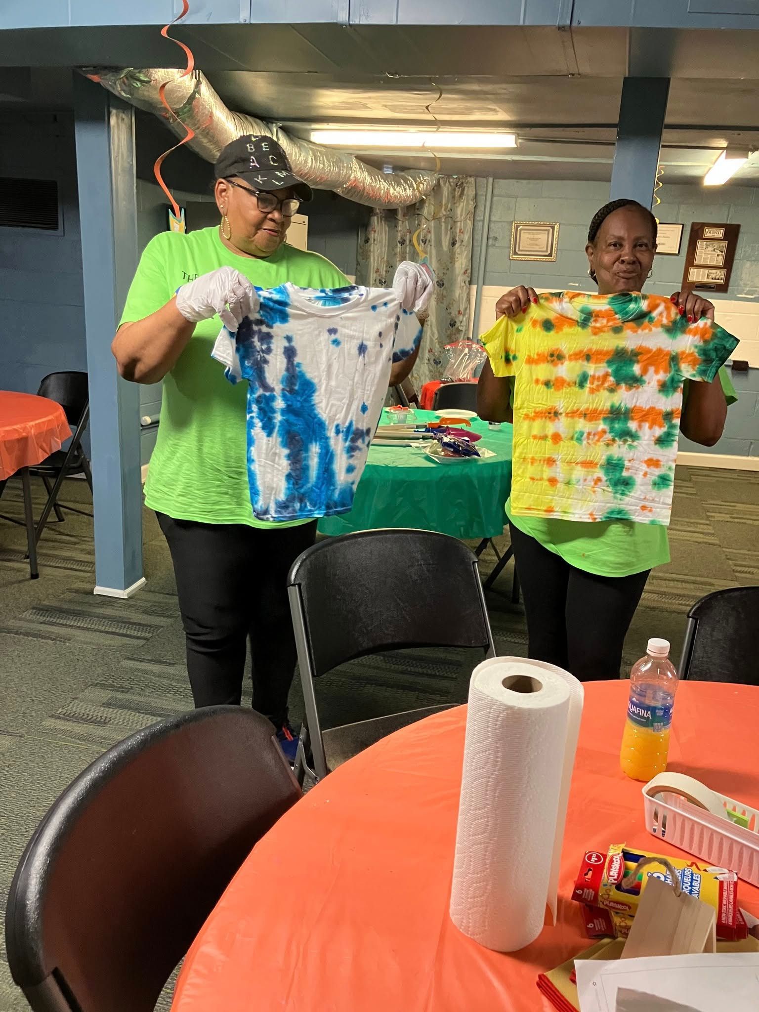 Two people in green shirts display their tie-dyed shirts in a room with orange-covered tables.