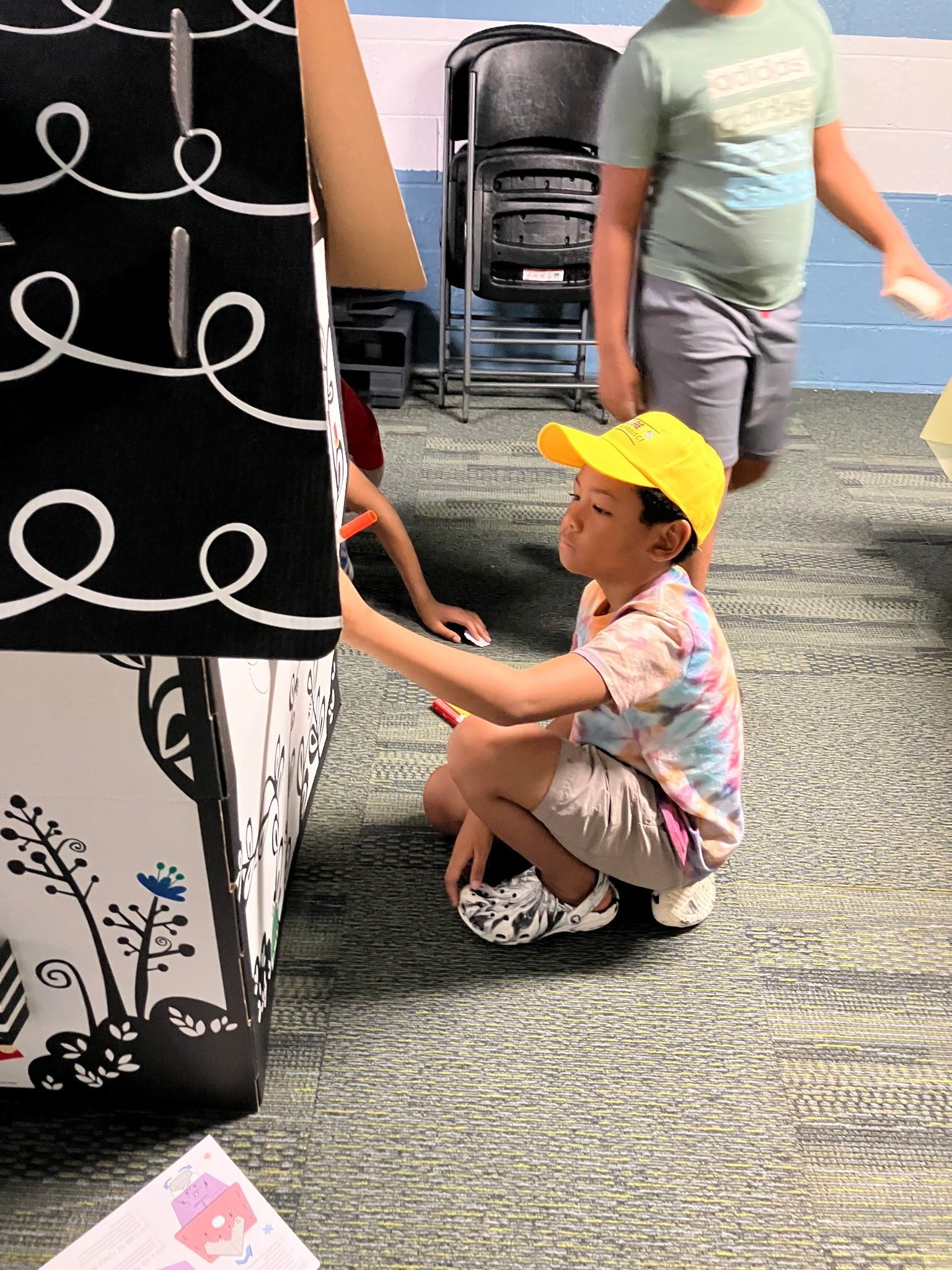 A child wearing a yellow cap and patterned shirt kneels while touching a decorated black-and-white display in a room.