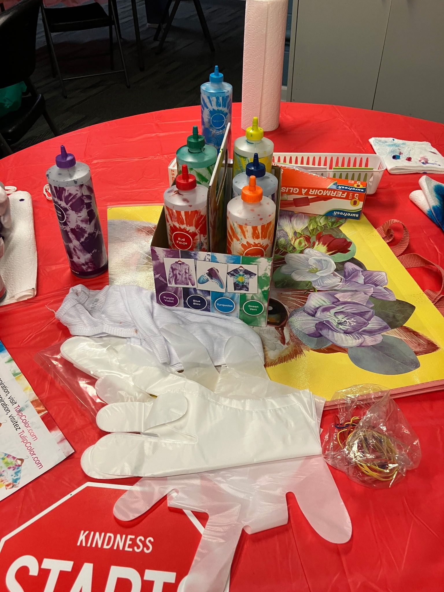 Craft supplies for tie-dyeing, including squeeze bottles, plastic gloves, and paper towels on a red tablecloth.